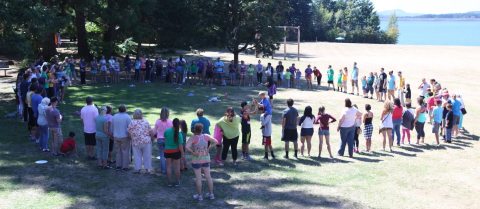 A large group of Courageous Kids Summer Camp attendees form a circle on a field next to a large body of water