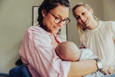 A mother holding a baby and a caregiver smiling nearby