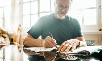 An older man sits at a table in a well lit room working on a crossword puzzle in the newspaper
