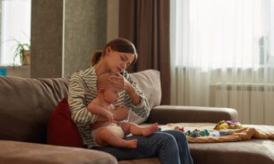 Young woman on couch feels forehead of baby on her lap while making a phone call.
