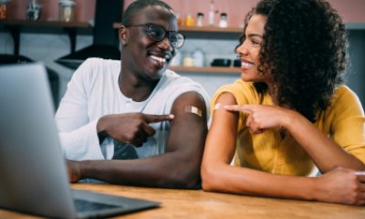 Two adults seated at a table in front of laptop point to arms, showing where they each received a vaccination