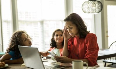 A stressed woman looking at her laptop screen rubs her head as her daughters ask her questions