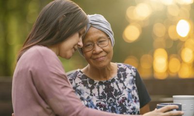 Two women outdoors -- one young and one older -- embrace and rest their for foreheads against each other. The older woman wears a headwrap indicating hair loss from cancer treatment.