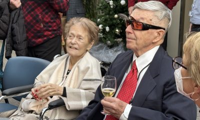 An elderly couple sits side by side at a reception. They wear formal clothing and hold glasses of cider in their hands.