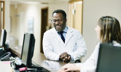 A smiling Black doctor leans on the counter of a nurses' station with a woman doctor