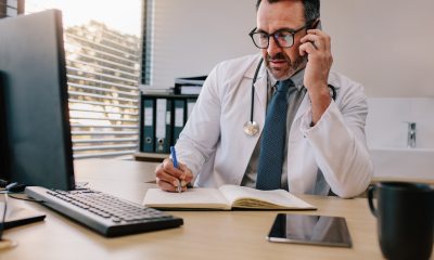 A healthcare provider at a desk and computer writes in a journal while speaking on the phone