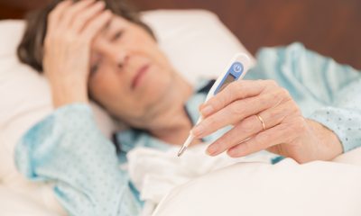 An older lady laying down takes her tempaerature and rests hand on her forehead.