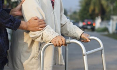 An older person walks across the street with a walker and the aid of someone off camera