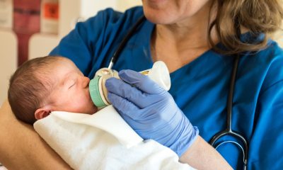 A gloved nurse in scrubs smiles and feeds a newborn baby with a bottle
