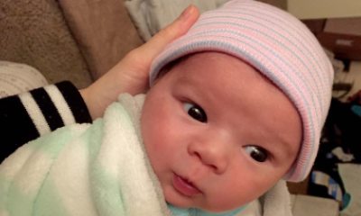 A wide-eyed newborn baby with a pink and blue striped beanie being cradled by an adult