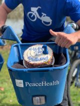 A PeaceHealth employee riding a bicycle with a cake in the basket, spelling out "Happy 1st Birthday"