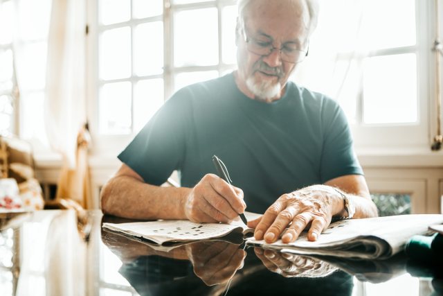 An older man sits at a table in a well lit room working on a crossword puzzle in the newspaper