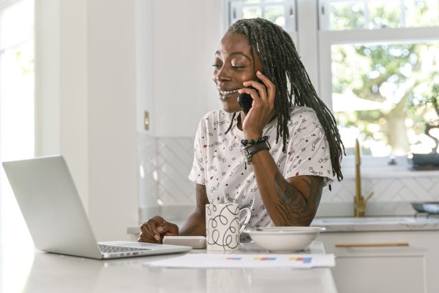 Woman looking at a laptop and holding a phone to her ear.