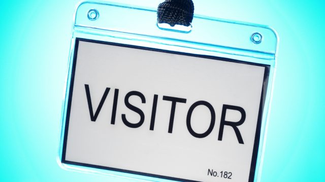A badge attached to a lanyard with the word "Visitor" in the foreground, as a group of silhouetted people stand in line in the background.
