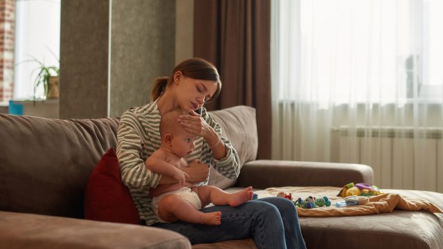 Young woman on couch feels forehead of baby on her lap while making a phone call.