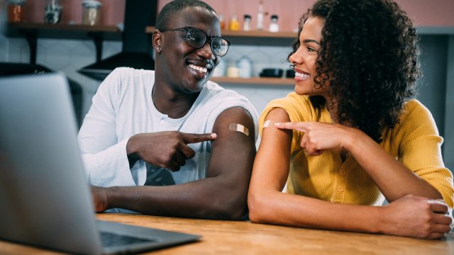 Two adults seated at a table in front of laptop point to arms, showing where they each received a vaccination