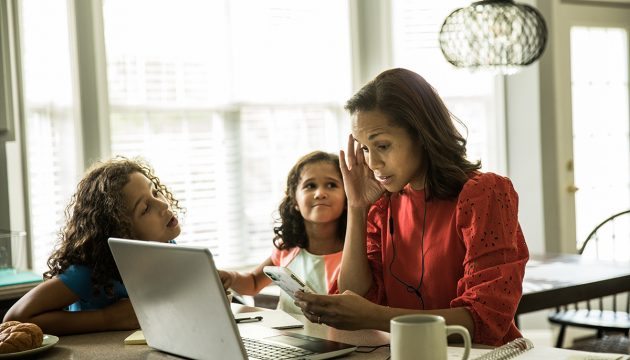 A stressed woman looking at her laptop screen rubs her head as her daughters ask her questions