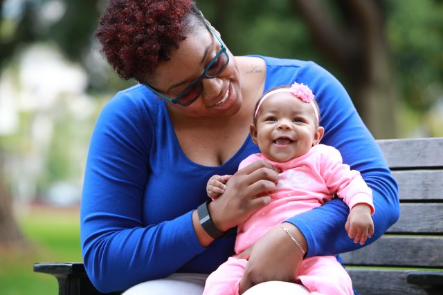 A smiling mother and newborn baby sit outside on a bench