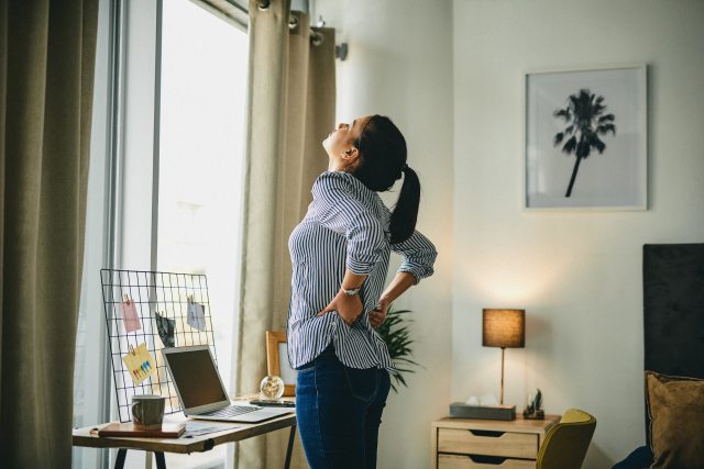 Adult woman stands in front of a desk, stretching her back.