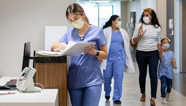 One woman provider reviews chart as another provider walks with two patients in clinic hallway.