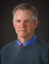 Headshot of man wearing shirt, tie and crewneck sweater