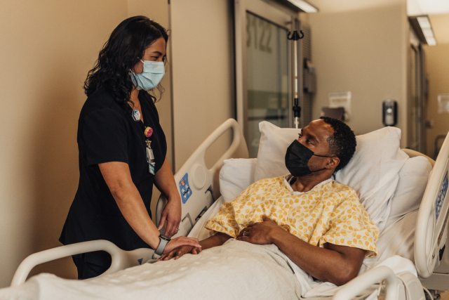 A PeaceHealth nurse stands bedside next to a patient lying in a hospital bed.