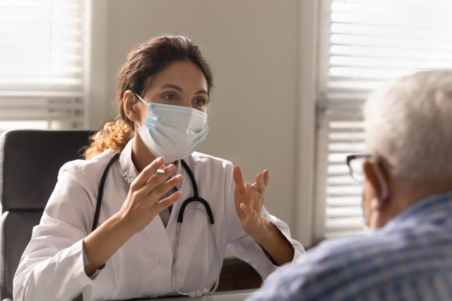 Close up of a female doctor wearing a face mask consulting with an older adult male patient  in doctor's office
