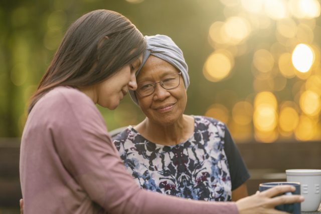 Two women outdoors -- one young and one older -- embrace and rest their for foreheads against each other. The older woman wears a headwrap indicating hair loss from cancer treatment.