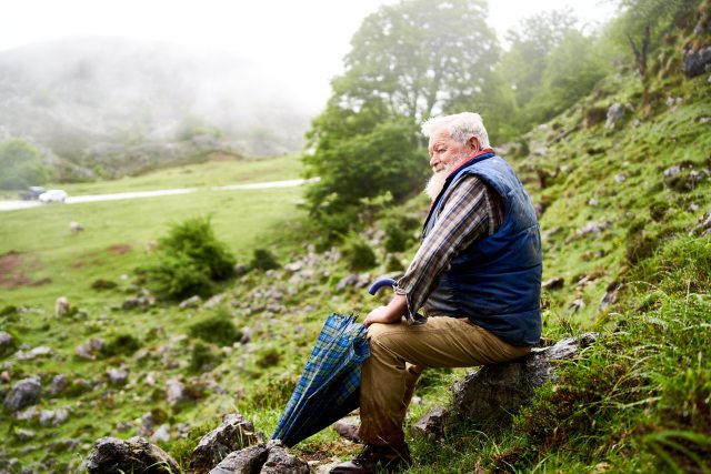 White-haired man sits on a rock on a hillside