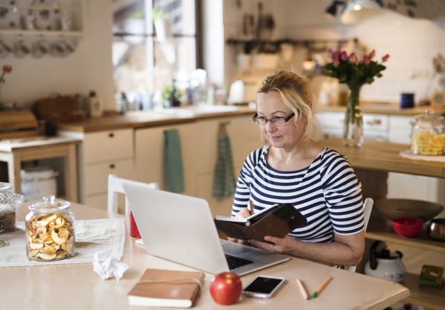 Older woman sits at kitchen table, looking at laptop screen and paper calendar