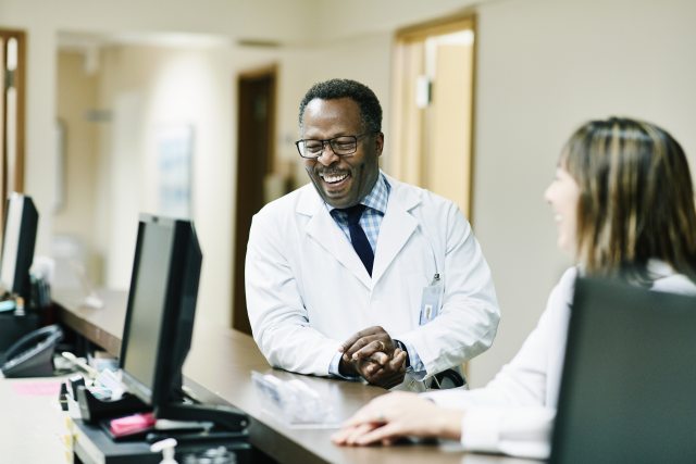 A smiling Black doctor leans on the counter of a nurses' station with a woman doctor