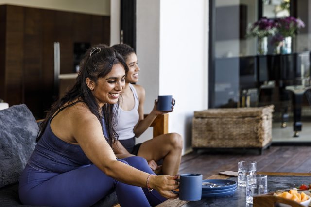 2 women with light brown skin relax on a couch with beverages