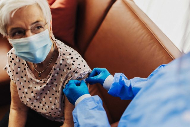 White haired woman in face mask has bandage applied to her arm by a healthcare provider in blue gloves and surgical gown after getting a shot or vaccine 