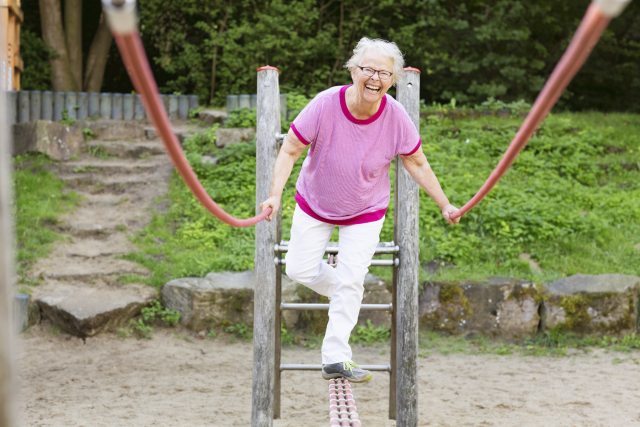 Smiling woman with short white hair balances on a piece of outdoor playground equipment
