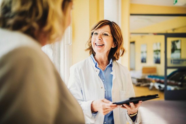 Female patient facing away from the camera talking to a  female healthcare provider in a lab coat holding a clipboard. 