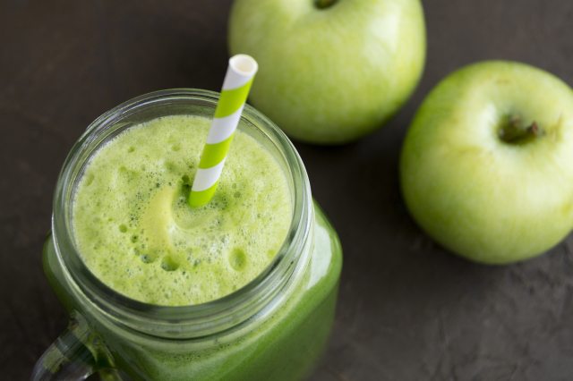 A green smoothie with a straw in a mason jar and green apples in the background