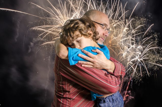 Father hugs young son while a fireworks display goes on in the background