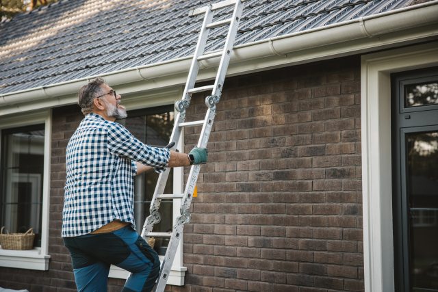 Man climbing up a ladder to clean the gutters on his house.