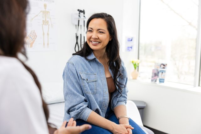 Woman with long dark hair smiles at a woman doctor wearing a white coat in an exam room