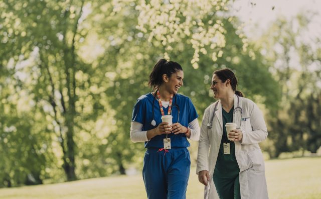 Two PeaceHealth Caregivers walk, talk and smile at each other in a park like setting.