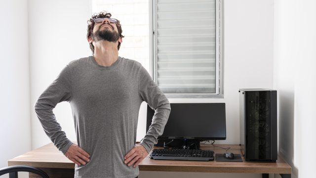 Bearded man in long-sleeve gray shirt looks up to ceiling, stretching back in front of desk