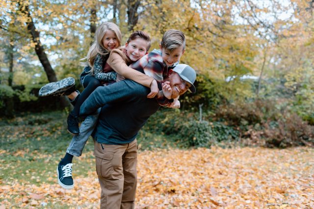 Three young children ride piggyback on a smiling red-haired man outside with fall leaves on the ground 
