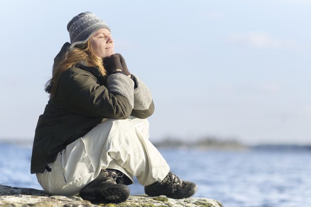 Photograph of adult white woman in knit hat and parka sitting cross-legged on a beach and meditating.