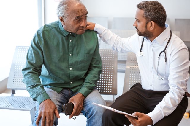 Bearded doctor sits and talks with older man in green button-up shirt