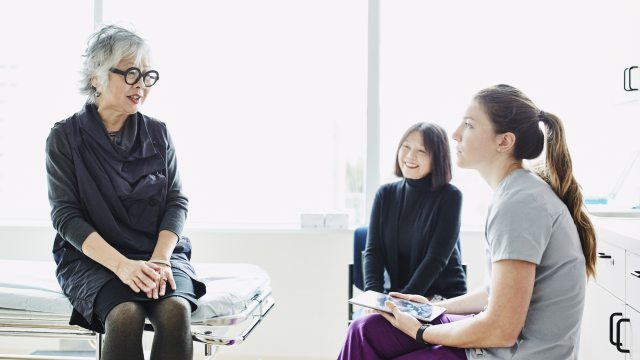 gray-haired woman on an exam table talks with a healthcare provider and dark-haired woman