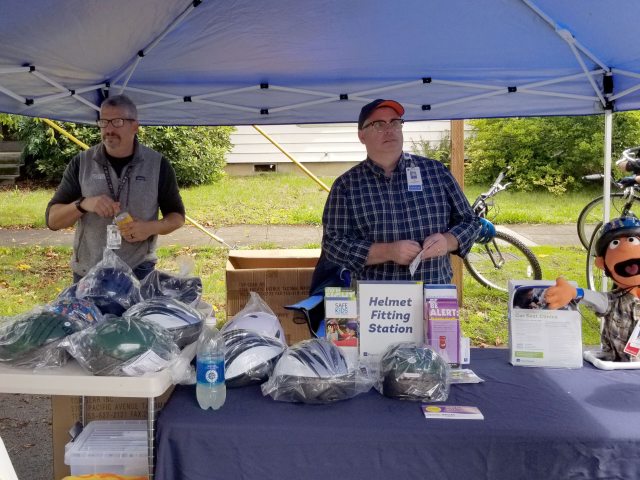 Two men stand behind a table filled with bicycle helmets.
