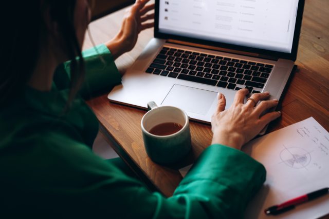 Woman at wooden table in green top viewing laptop screen. A green coffee cup and notepad are on the table next to her hands.
