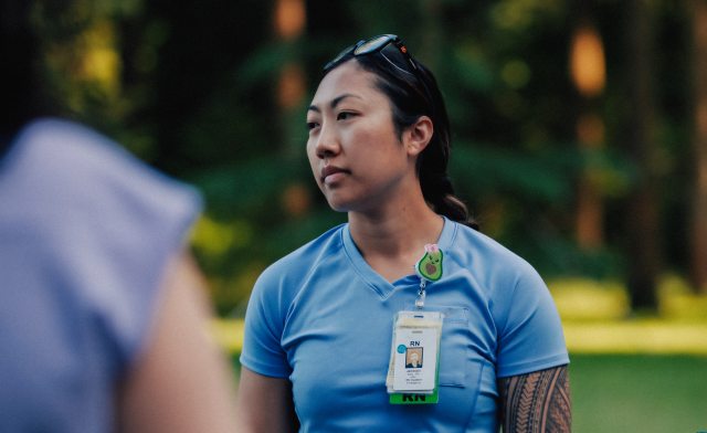 A badged nurse standing in a park-like setting looking at something or someone off camera.