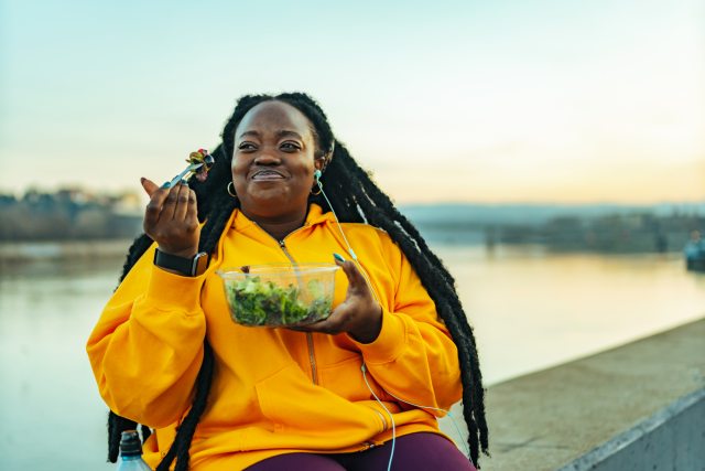 Woman wearing a yellow sweatshirt enjoying a salad near water.