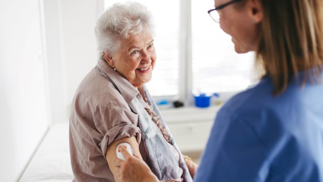 Woman with white hair smiles at caregiver applying a glucose monitor to arm